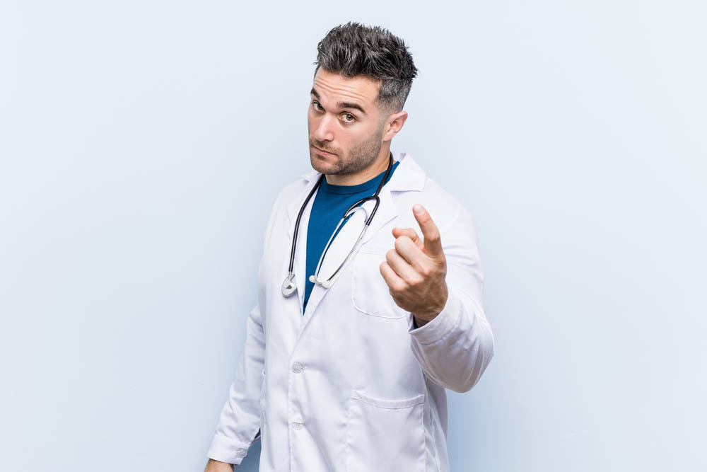 Young handsome doctor man pointing with finger at you as if inviting come closer.